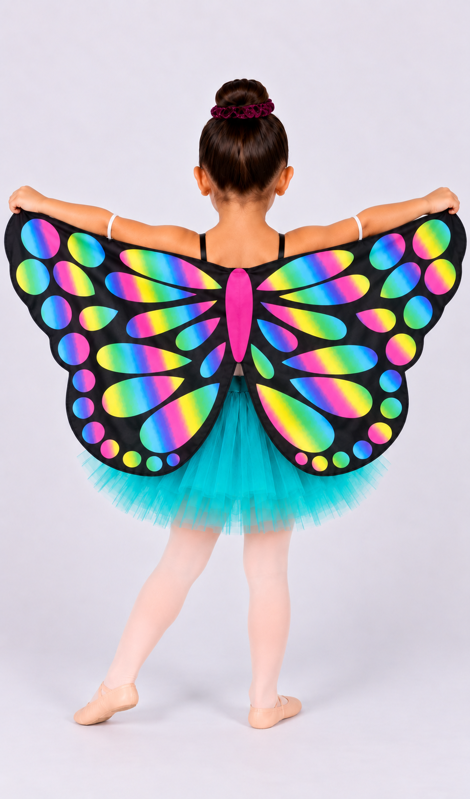 A young girl from behind wearing a black butterfly wing costume with a vibrant rainbow gradient pattern and a turquoise tutu skirt, posing against a light gray studio background.
