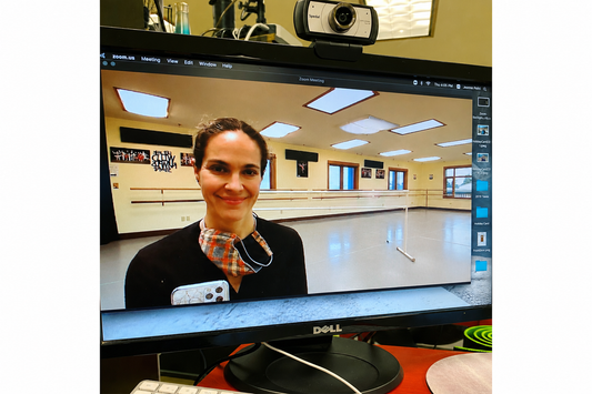  A computer monitor on a desk showing a video call screen with a woman in a dance studio, with a webcam mounted above the monitor.