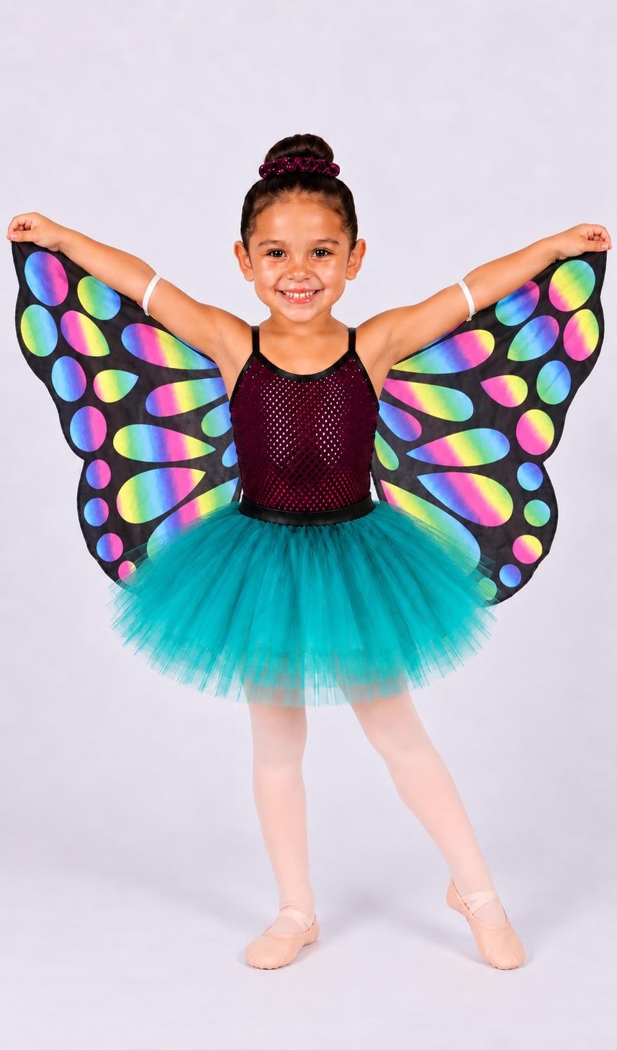 A young girl modeling a butterfly costume against a light gray studio background. The costume features a dark burgundy mesh bodysuit, a bright teal tulle tutu skirt, and large black wings with a vibrant rainbow pattern.